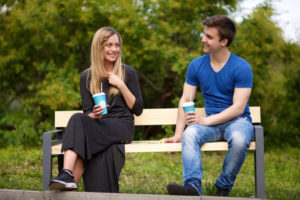 公園のベンチに座り飲み物を手に談笑する髪の長い黒い服を着た女性とデニムジーンズをはいて青色のTシャツをきた短髪の男性